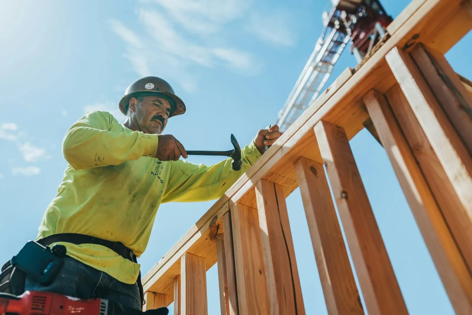 A framer in a hard hat and hi-vis shirt nailing a wall frame on a sunny job site
